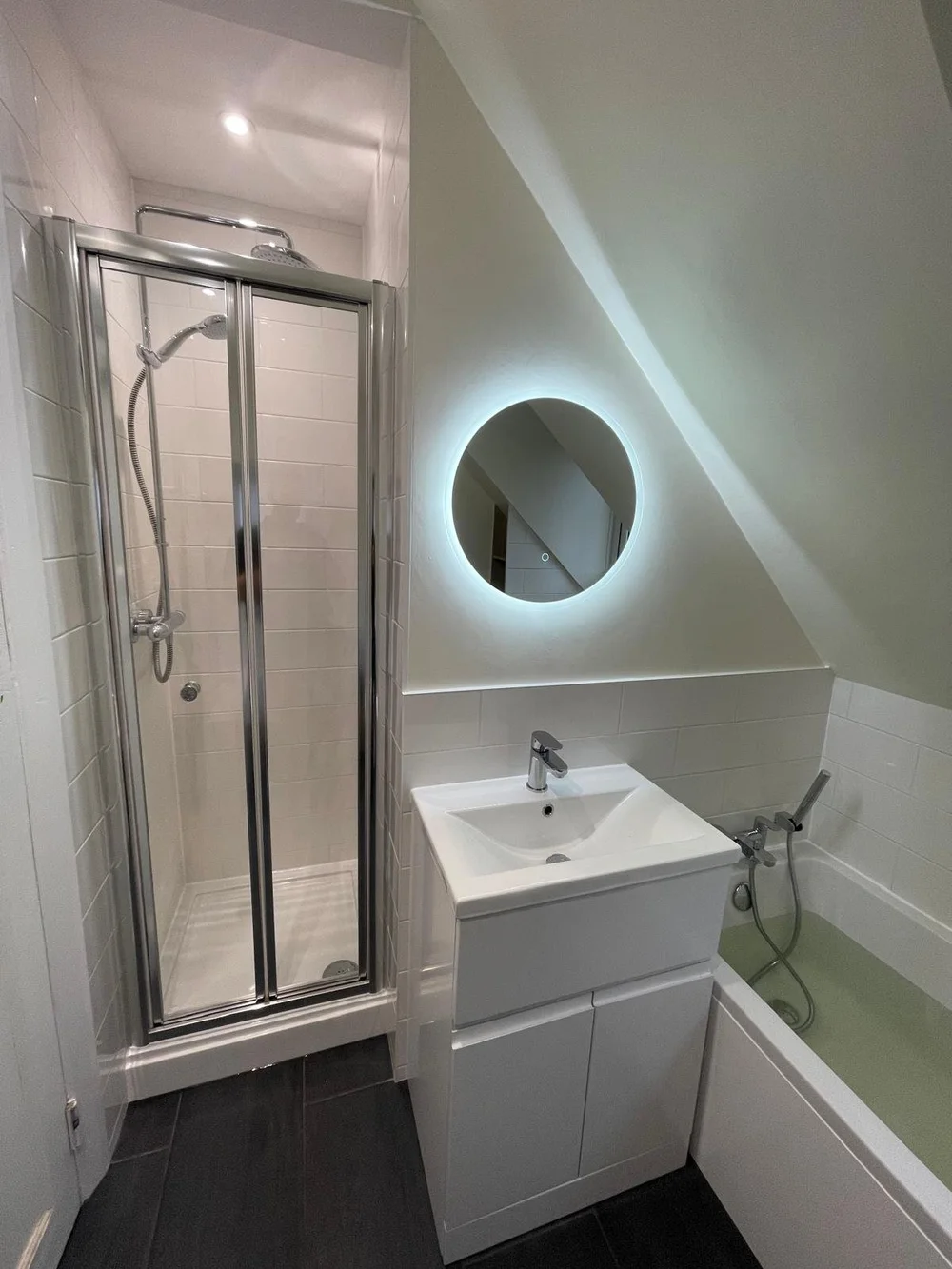 Compact loft shower room with backlit round mirror and bi-fold shower door — bathroom renovation Woking