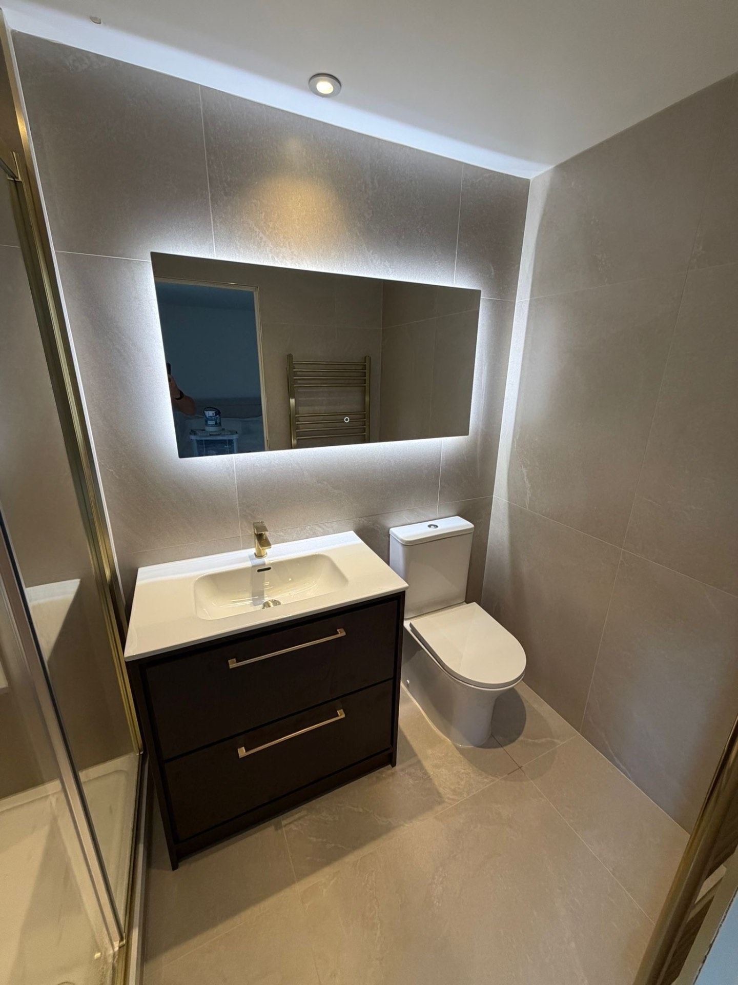 Ensuite with backlit mirror, dark wood vanity and brushed gold shower enclosure — bathroom installation Woking