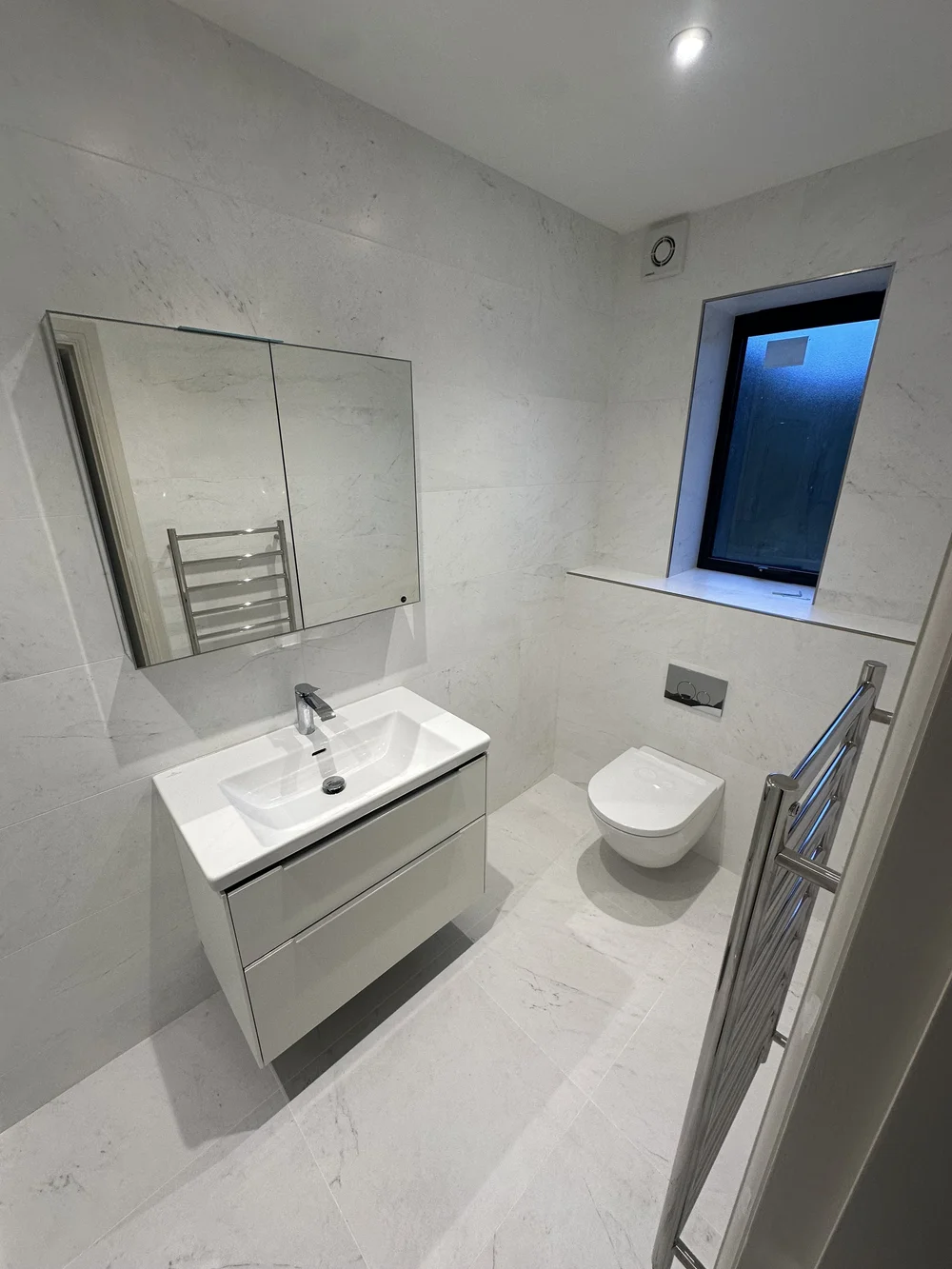 White marble ensuite with wall-hung vanity, mirrored cabinet and back-to-wall WC — Woking