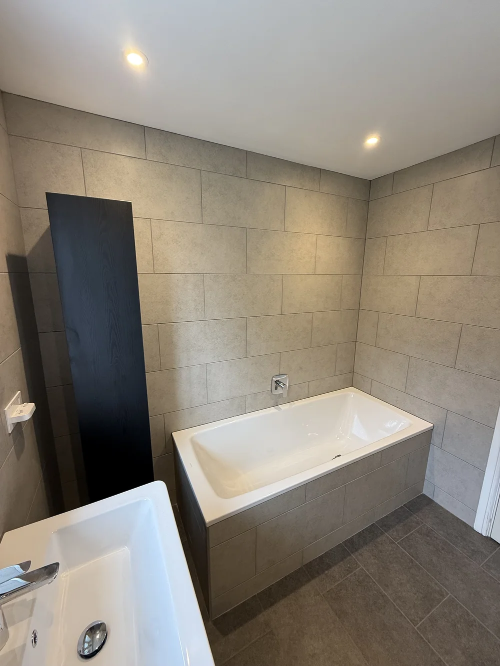 Stone-tiled bathroom with panelled bath and tall black storage column — bathroom renovation Woking