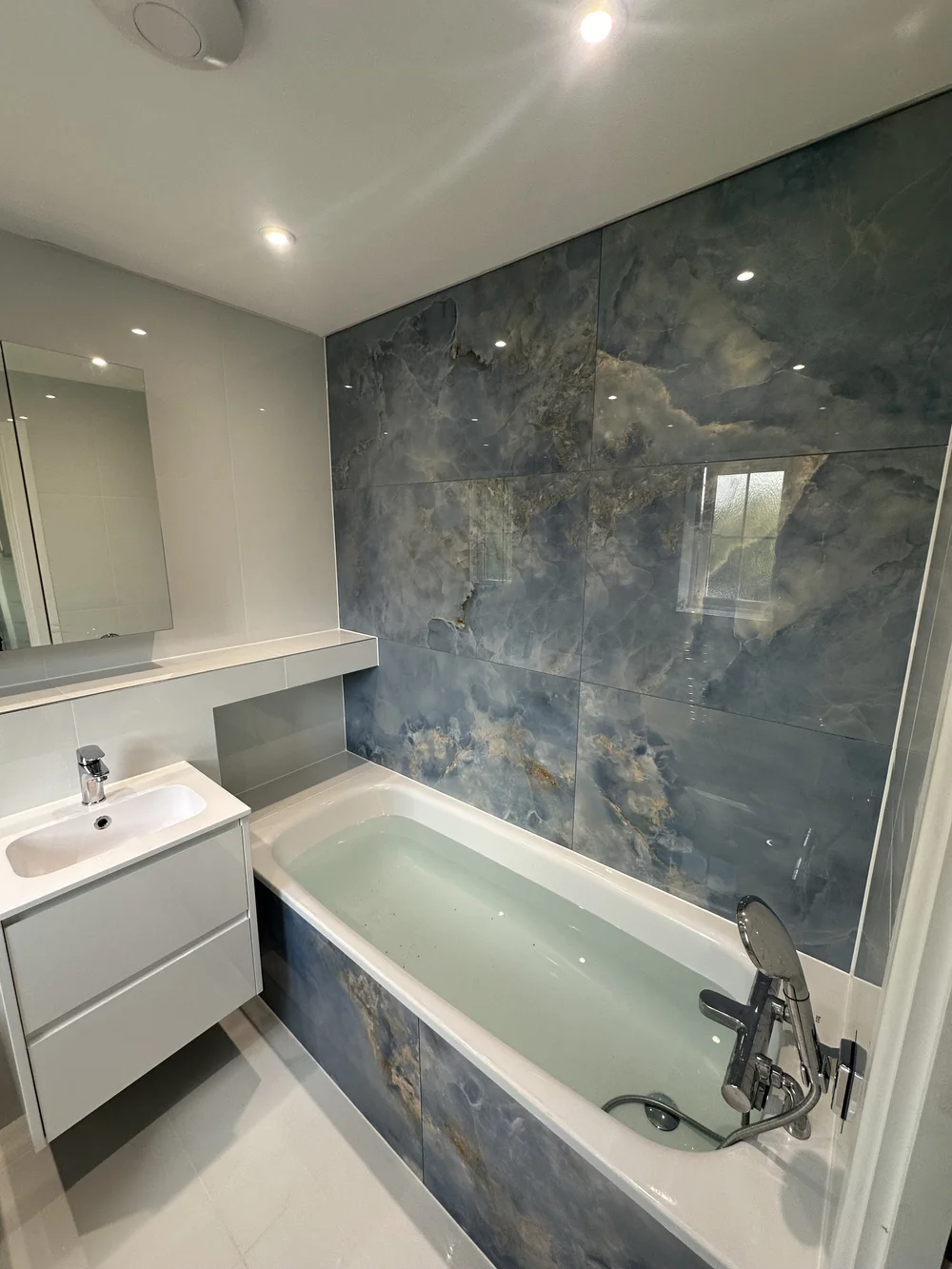 Family bathroom with blue onyx-effect feature wall, panelled bath and floating white vanity — bathroom installation Woking